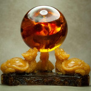 Puede incluir: Una esfera decorativa de color ámbar, sostenida por tres figuras con forma de dragón sobre una base marrón oscuro. La esfera tiene una superficie reflectante y está llena de detalles internos. Las figuras son de color amarillo dorado.