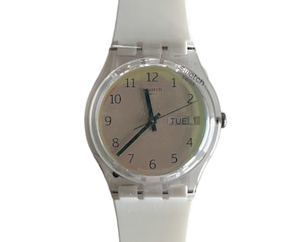 Peut inclure: Une montre-bracelet en plastique blanc avec un cadran transparent et des aiguilles noires. Le cadran de la montre comporte des chiffres noirs et un affichage du jour de la semaine qui indique "TUE 1".