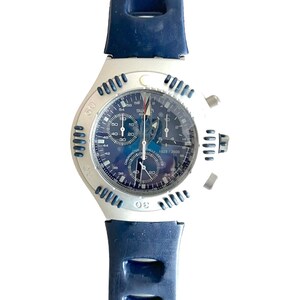 May include: A silver and blue chronograph wristwatch with a blue face and a black rubber strap. The watch has a date window and a tachymeter scale.