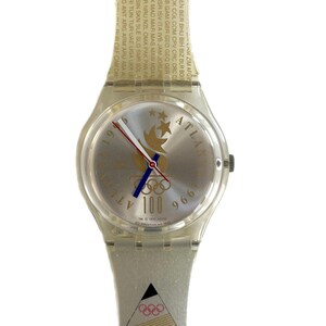 Puede incluir: Reloj Swatch transparente con esfera plateada con el logotipo de los Juegos Olímpicos de Atlanta 1996. La correa del reloj es transparente con texto y los anillos olímpicos. El reloj tiene detalles en rojo, azul y dorado.