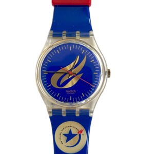Puede incluir: Un reloj Swatch azul y plateado con un diseño de llama dorada en la esfera. El reloj tiene una correa de plástico azul con un acento rojo y un logotipo de estrella blanca en la parte inferior.