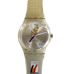 Puede incluir: Un reloj Swatch plateado con una esfera blanca y detalles dorados. La esfera presenta los anillos olímpicos y el texto "Atlanta 1996" y "100". La correa es de plástico transparente con texto dorado y los anillos olímpicos.