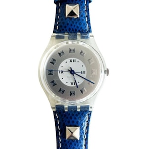 May include: A clear plastic wristwatch with a white face and silver Roman numeral hour markers. The watch has a blue leather band with silver studs.