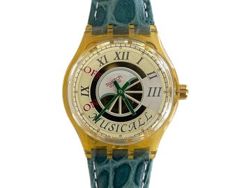 Peut inclure: Une montre Swatch dorée avec un cadran crème et des chiffres romains. Le cadran présente un motif musical avec des aiguilles vertes et le mot "MUSICALLL". Elle a un bracelet en cuir vert d'eau.