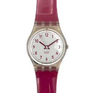 Puede incluir: Un reloj de pulsera analógico blanco con una caja plateada y una correa de plástico rosa. La esfera del reloj tiene agujas de hora y minuto negras y una aguja de segundos roja. La esfera del reloj tiene números negros del 1 al 12 y marcas negras para las otras horas. La esfera del reloj también tiene la palabra "swatch" en letras negras y la palabra "swiss" en letras negras más pequeñas.