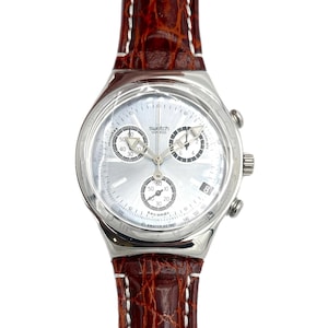May include: A silver Swatch Swiss chronograph watch with a light-colored face and three sub-dials. The watch has a brown leather band with white stitching. The text "Swatch Swiss" and "SWATCH AG 1997" are visible on the watch face.