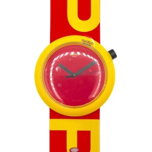 Op de afbeelding: Een Swatch Swiss horloge met een felrode wijzerplaat en zwarte wijzers, gevat in een gele behuizing. Het horloge is bevestigd aan een rode en gele band met het woord "POP" in grote gele letters. Het horloge heeft een zwarte kroon.