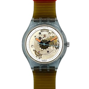 May include: A transparent Swatch watch with a skeleton dial, showcasing the inner workings. The watch has a silver case, a brown strap, and a red band. The watch face displays the Swatch logo.