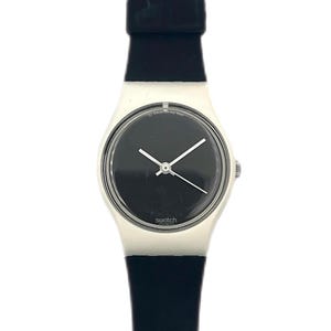 May include: A Swatch watch with a black dial and white hands. The watch has a white case and a black rubber strap. The word "swatch" is printed on the watch face. The watch is a classic, minimalist design.