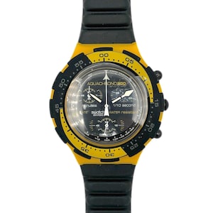 Op de afbeelding: Zwart en gele Swatch Aquachrono 200 polshorloge. Het horloge heeft een zwarte wijzerplaat met witte wijzers en markeringen, een gele lunette en een zwarte rubberen band. Het is waterdicht en heeft een 1/10 seconde timer.