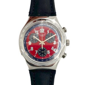 May include: A Swatch chronograph watch with a red dial, silver case, and black leather band. The watch face features black sub-dials, white numerals, and the Swatch logo. The watch has a date display at the 3 o'clock position.