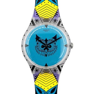 Op de afbeelding: Een Swatch Swiss horloge met een blauwe wijzerplaat met een zwart uilontwerp. De horlogeband heeft een kleurrijk geometrisch patroon met gele, blauwe en zwarte accenten. De horlogekast is zilver.