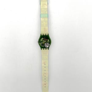 May include: A transparent Swatch watch with a green face and a clear band. The watch face reveals the internal mechanisms and a silver battery. The band has a repeating pattern of small, colorful images.