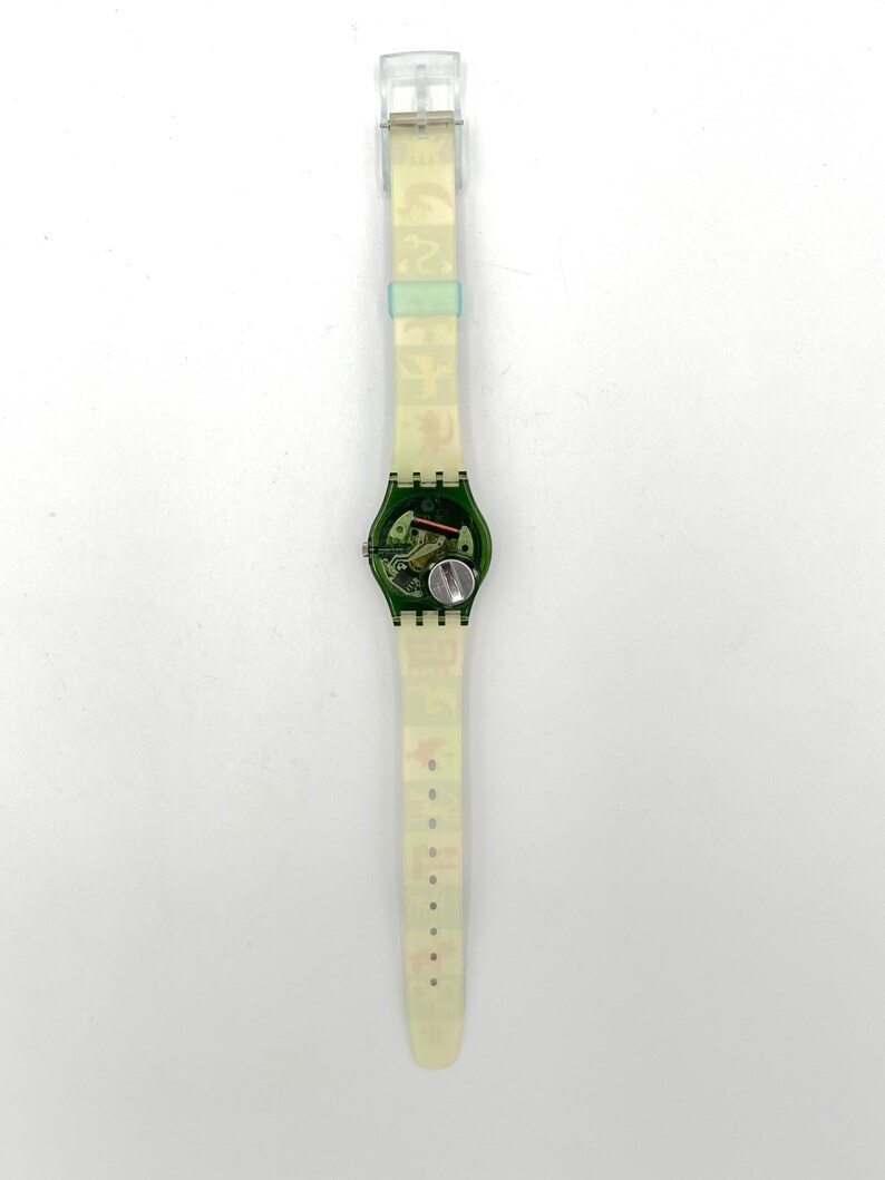 May include: A transparent Swatch watch with a green face and a clear band. The watch face reveals the internal mechanisms and a silver battery. The band has a repeating pattern of small, colorful images.