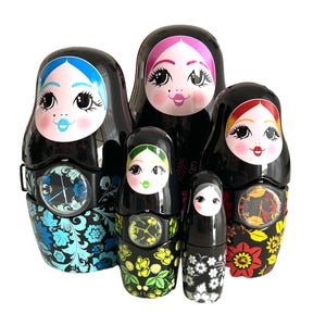 Pode incluir: Um conjunto de cinco bonecas russas pretas com rostos pintados e mostradores de relógio florais. As bonecas têm cabelos de cores diferentes e padrões florais em suas roupas. A boneca maior tem cabelo azul e um padrão floral azul.