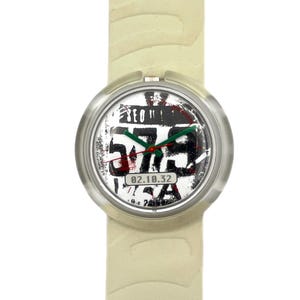 Puede incluir: Un reloj de pulsera con una correa color crema y una caja plateada. La esfera del reloj presenta un diseño en blanco y negro con los números "575" en negrita, y el texto "SEO!" y "USA". La fecha "02.10.32" también es visible.
