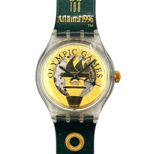 Puede incluir: Reloj Swatch de los Juegos Olímpicos de Atlanta 1996. El reloj tiene una caja de plástico transparente, una esfera dorada con los anillos olímpicos y la inscripción "Olympic Games". La correa es verde oscuro con detalles dorados y la inscripción "Atlanta 1996".