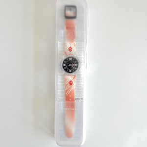 Puede incluir: Un reloj de pulsera con estampado blanco y rojo, esfera negra y agujas plateadas. El reloj est&aacute; en una caja de pl&aacute;stico transparente con la palabra "Swatch" impresa en el lateral.
