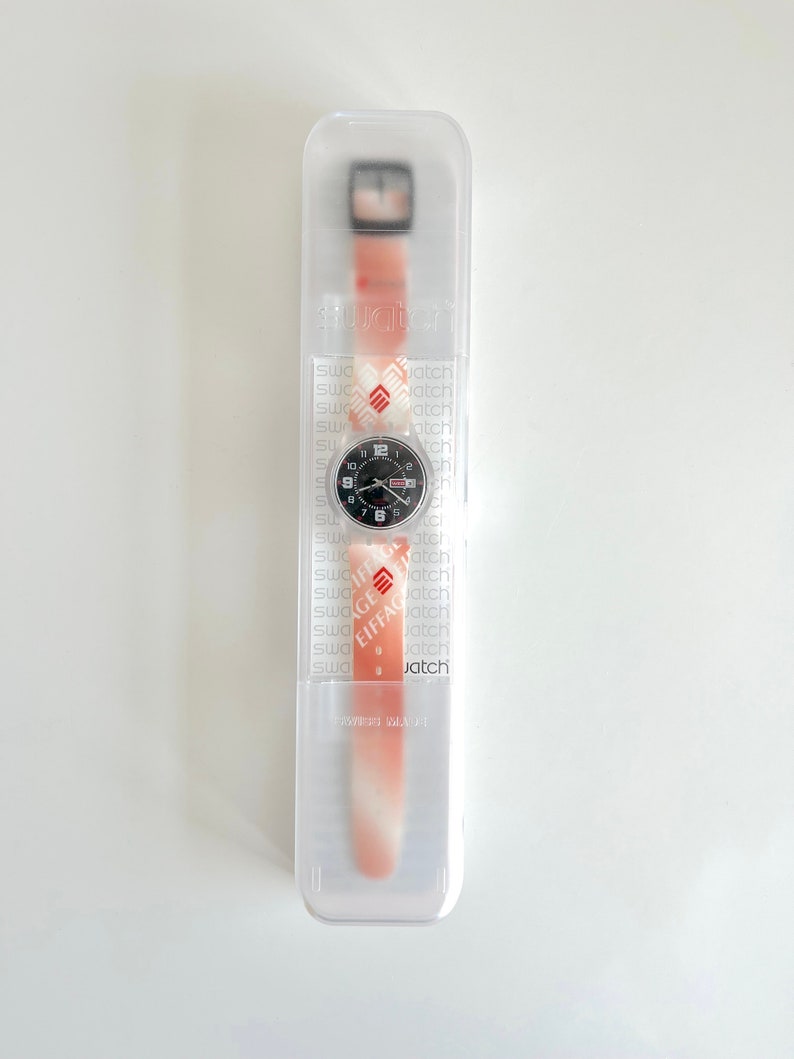 Puede incluir: Un reloj de pulsera con estampado blanco y rojo, esfera negra y agujas plateadas. El reloj est&aacute; en una caja de pl&aacute;stico transparente con la palabra "Swatch" impresa en el lateral.
