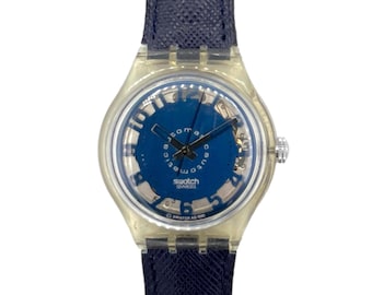 May include: A Swatch automatic watch with a transparent plastic case and a dark blue dial. The dial features white numerals and hands, with the brand name visible. The watch has a dark blue leather strap.