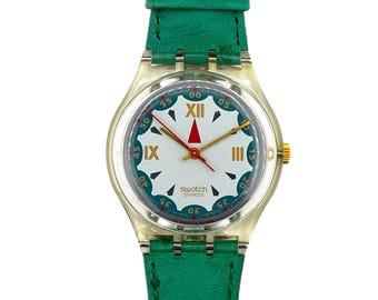 Peut inclure: Une montre Swatch avec un boîtier en plastique transparent et un cadran blanc avec des accents dorés et noirs. La montre a une trotteuse rouge et des aiguilles des heures et des minutes dorées. Le bracelet en cuir vert complète le cadran.