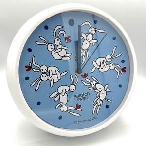 Op de afbeelding: Een witte Swatch-klok met een lichtblauwe wijzerplaat met cartoonkonijnen en rode bloemaccenten. De klok heeft zwarte wijzers en de tekst "Swatch Swiss" en "Swatch AG 2003".
