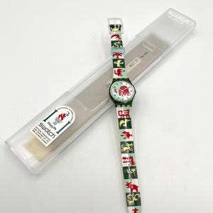 May include: A Swatch Quartz watch with a green and white band featuring red and green graphic designs. The watch face is green with red accents and the number markers. The watch is in its original clear plastic case, with the text "Swatch Quartz" on the case.