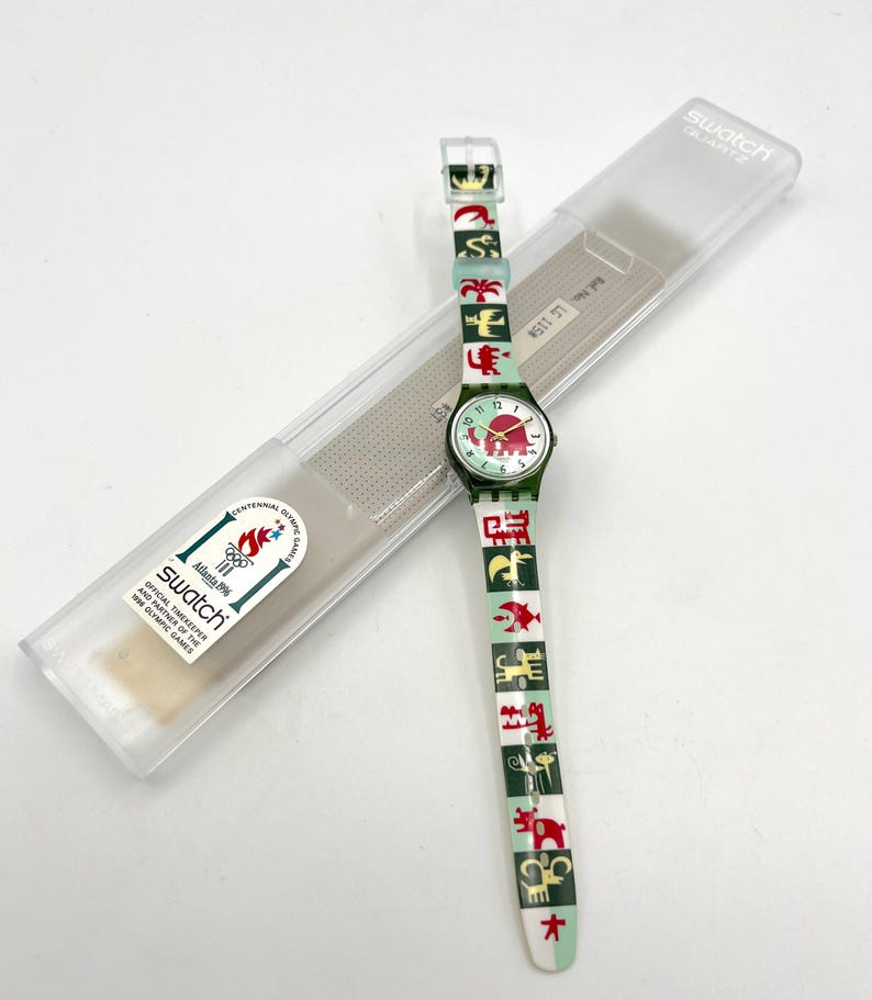 May include: A Swatch Quartz watch with a green and white band featuring red and green graphic designs. The watch face is green with red accents and the number markers. The watch is in its original clear plastic case, with the text "Swatch Quartz" on the case.