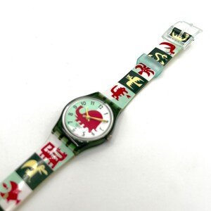 May include: A vintage wristwatch with a green case and a light green face featuring a red elephant design. The watch band is white and green with repeating red and gold animal and symbol patterns. The watch has gold hands and a clear plastic buckle.