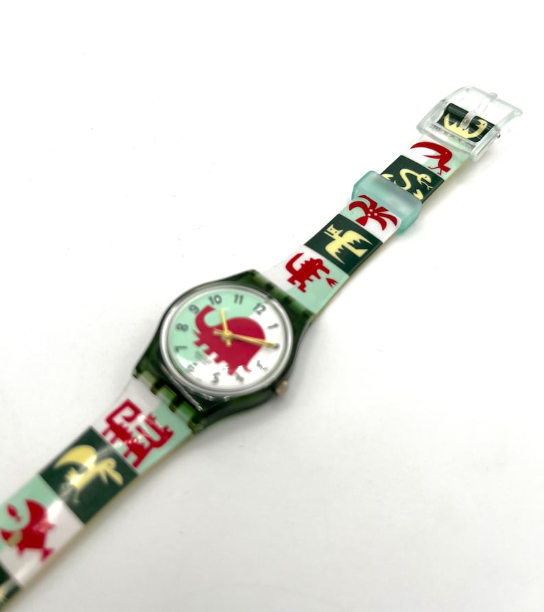 May include: A vintage wristwatch with a green case and a light green face featuring a red elephant design. The watch band is white and green with repeating red and gold animal and symbol patterns. The watch has gold hands and a clear plastic buckle.