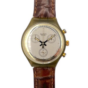 以下が含まれることがあります： 明るい文字盤、ゴールドトーンのアクセント、茶色の革バンドが特徴のヴィンテージスウォッチスイスウォッチ。時計には3つのサブダイヤルとスウォッチのロゴが付いています。時計ケースはオリーブグリーンです。クラシックな時計。