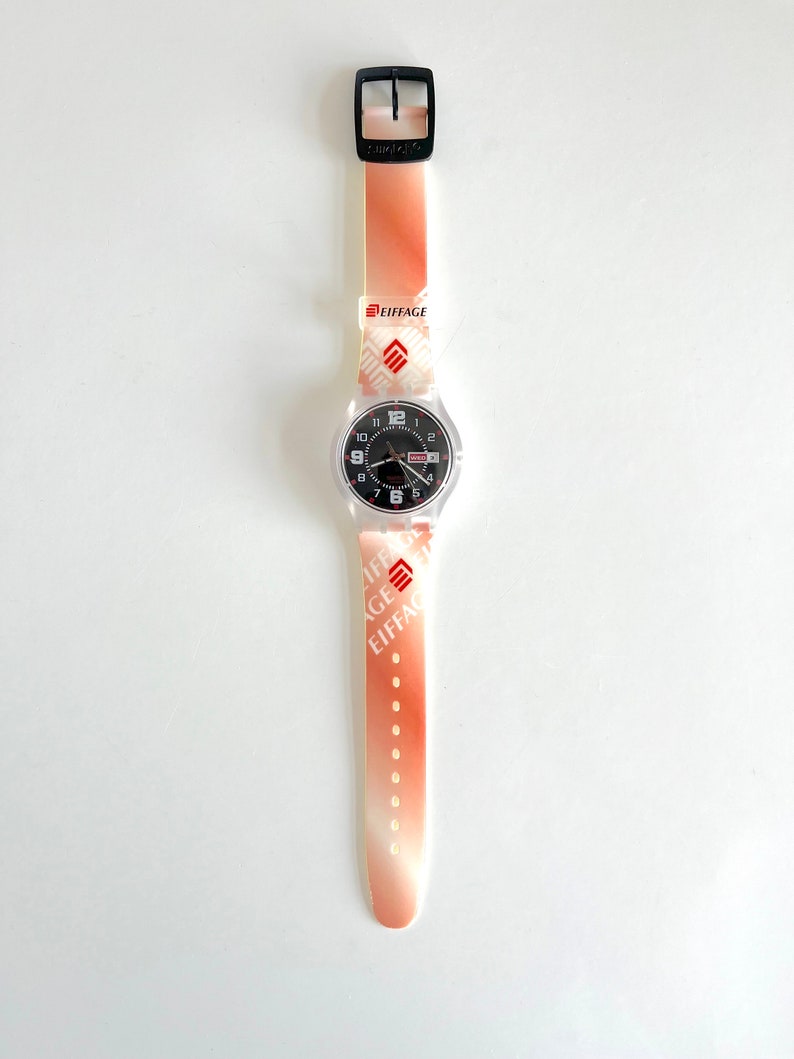 Puede incluir: Un reloj Swatch blanco y naranja con una esfera negra y un logotipo "Eiffage" rojo en la correa.