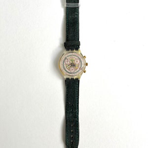 May include: A gold-toned wristwatch with a white face and black hands. The watch has a green corduroy strap and a clear plastic case. The watch face features a chronograph with three sub-dials and the text "Swatch" printed on it.