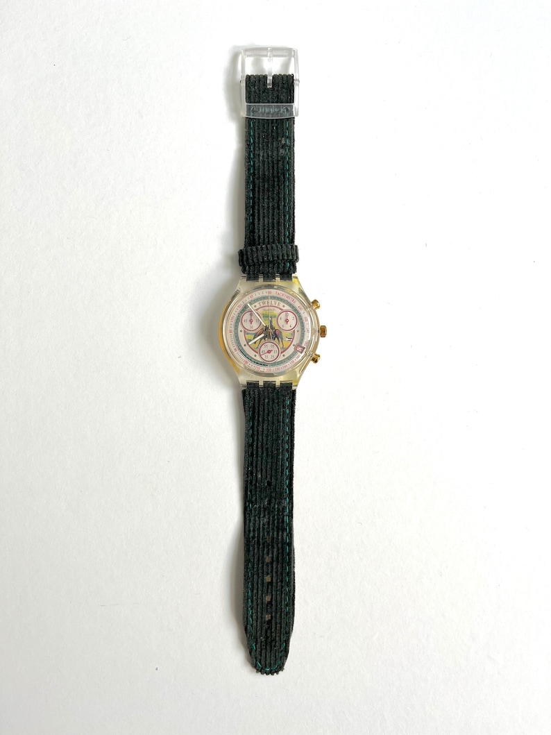 May include: A gold-toned wristwatch with a white face and black hands. The watch has a green corduroy strap and a clear plastic case. The watch face features a chronograph with three sub-dials and the text "Swatch" printed on it.