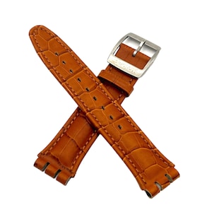 Op de afbeelding: Een paar bruin lederen horlogebandjes met een krokodillenleertextuur. De bandjes zijn gekruist, met een zilverkleurige gesp waarop het woord "swatch" is gegraveerd. De bandjes zijn nieuw.