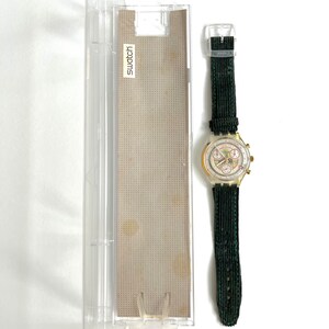 May include: A gold-toned Swatch chronograph wristwatch with a white face and black hands. The watch has a green fabric strap and comes in a clear plastic case with the word "Swatch" printed on it.