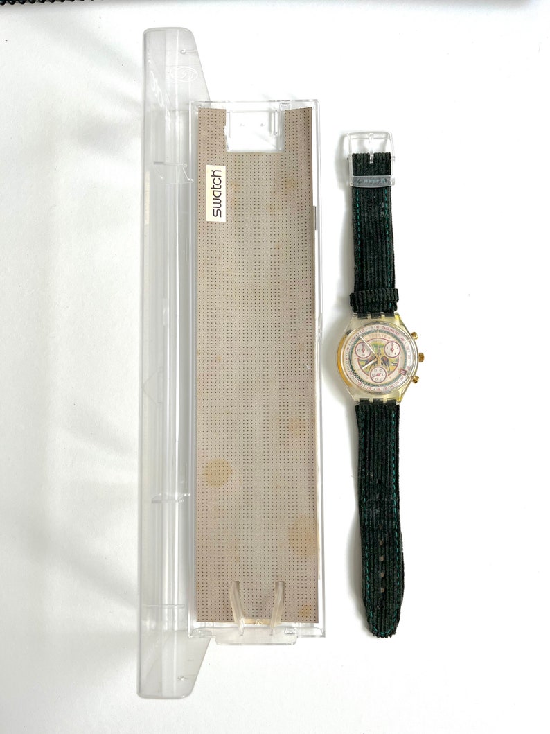 May include: A gold-toned Swatch chronograph wristwatch with a white face and black hands. The watch has a green fabric strap and comes in a clear plastic case with the word "Swatch" printed on it.