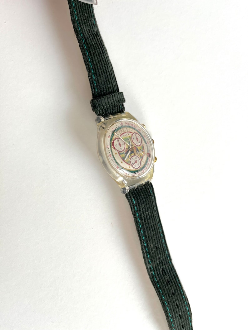 May include: A silver and white wristwatch with a black and green corduroy band. The watch face has a white background with black and red numbers and hands. The watch face also has three smaller dials with black and white numbers.