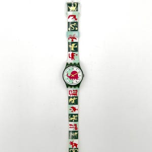 May include: A vintage wristwatch with a white face, black numbers, and red hands. The watch band is light green with red and yellow animal graphics. The watch case is dark green. The watch is a unique accessory.