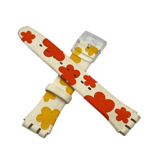 May include: Cream-colored watch band with a floral pattern in orange and yellow. The watch band has a clear plastic buckle and is designed to fit a watch. The floral design is repeated across the band.