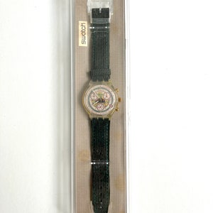 May include: A clear plastic watch with a white face and black hands. The watch has a black fabric band and is in a clear plastic box with the word "Swatch" printed on it. The box also says "Swiss Made".