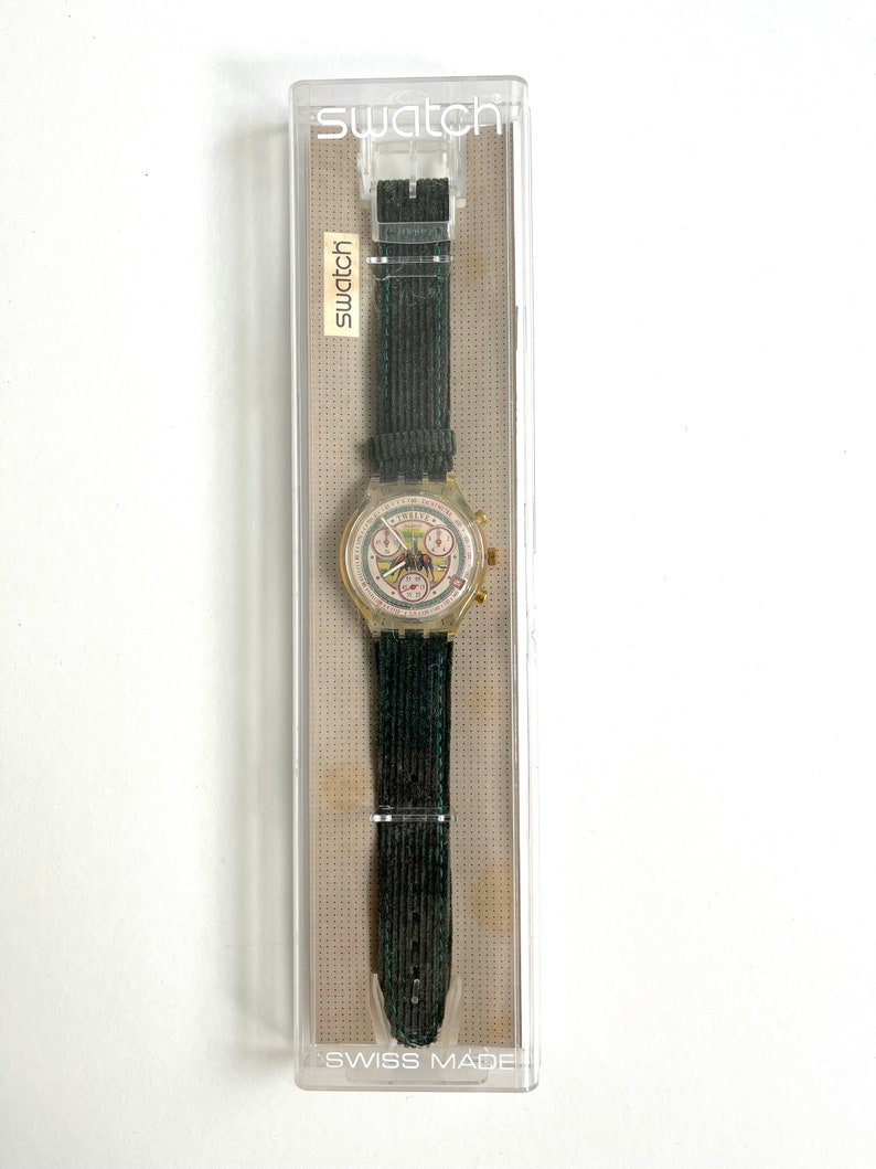 May include: A clear plastic watch with a white face and black hands. The watch has a black fabric band and is in a clear plastic box with the word "Swatch" printed on it. The box also says "Swiss Made".