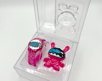 Puede incluir: Un juego de reloj y figura rosa en una caja de plástico transparente. El reloj y la figura presentan un diseño de personaje de dibujos animados con una cara azul y blanca. La correa del reloj y la figura tienen un patrón blanco.