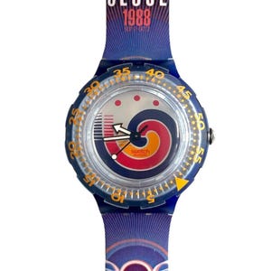 Puede incluir: Reloj Swatch azul con correa a juego, con el texto "SEOUL 1988". La esfera del reloj tiene un diseño colorido con detalles en rojo, naranja y azul. La correa del reloj tiene un patrón repetido de anillos olímpicos.