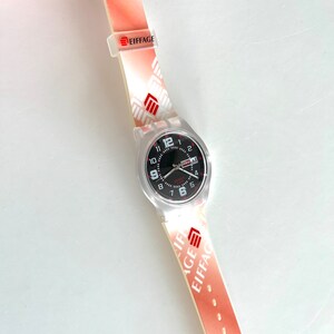 Puede incluir: Un reloj de pulsera blanco con una esfera negra y n&uacute;meros rojos. El reloj tiene una correa de pl&aacute;stico transparente con un patr&oacute;n rojo y blanco y la palabra "EIFFAGE" impresa en ella.