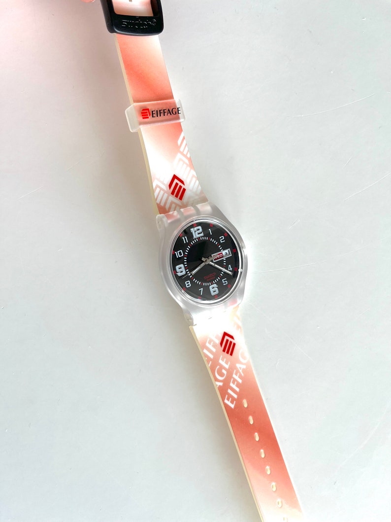 Puede incluir: Un reloj de pulsera blanco con una esfera negra y n&uacute;meros rojos. El reloj tiene una correa de pl&aacute;stico transparente con un patr&oacute;n rojo y blanco y la palabra "EIFFAGE" impresa en ella.