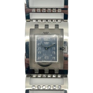 Op de afbeelding: Zilverkleurig Swatch-horloge met een rechthoekige wijzerplaat en bijpassende band. De wijzerplaat is donkerblauw met witte cijfers en wijzers. De band heeft heldere, vierkante kristallen. Een stijlvol accessoire, Zwitsers gemaakt.