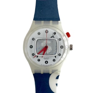Op de afbeelding: Een wit Swatch-horloge met een blauw-wit gepatenteerd bandje. De wijzerplaat van het horloge heeft een witte achtergrond met zwarte cijfers en een rode secondewijzer. De wijzerplaat heeft ook een klein vierkant venster met een grijs-wit patroon.
