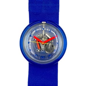 Puede incluir: Un reloj de pulsera azul con esfera transparente, que muestra los mecanismos internos. El reloj tiene una correa azul y agujas rojas. La esfera redonda está enmarcada en azul.