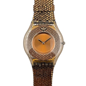 May include: A Swatch watch with a transparent case and a copper-colored dial. The watch features a brown, dotted band and a gold-colored bezel. The watch face has the word "Swatch" printed on it.